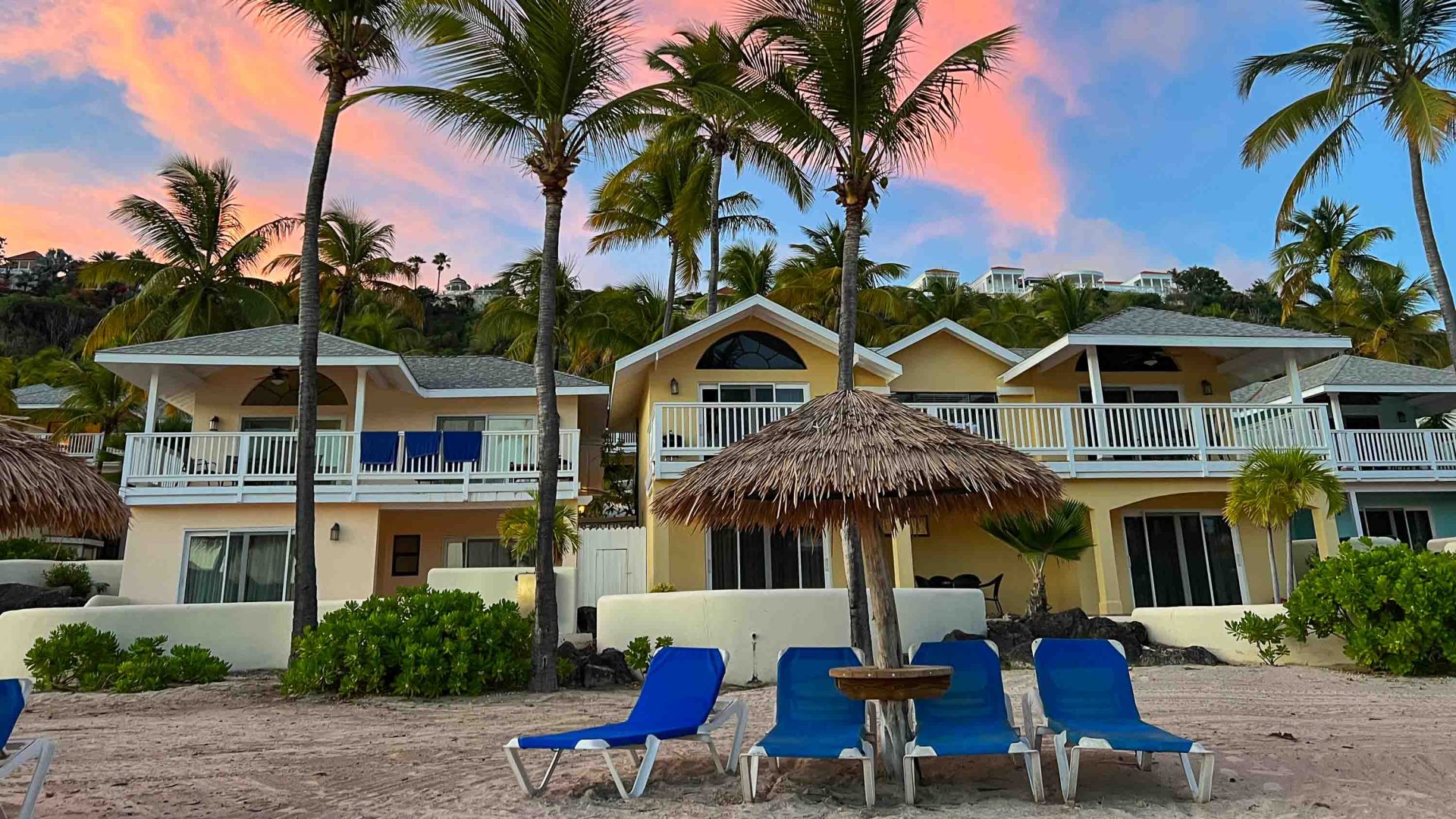 The resort where the writer's family stayed. It has cream coloured villas and palm trees. There is a pink sunset sky.