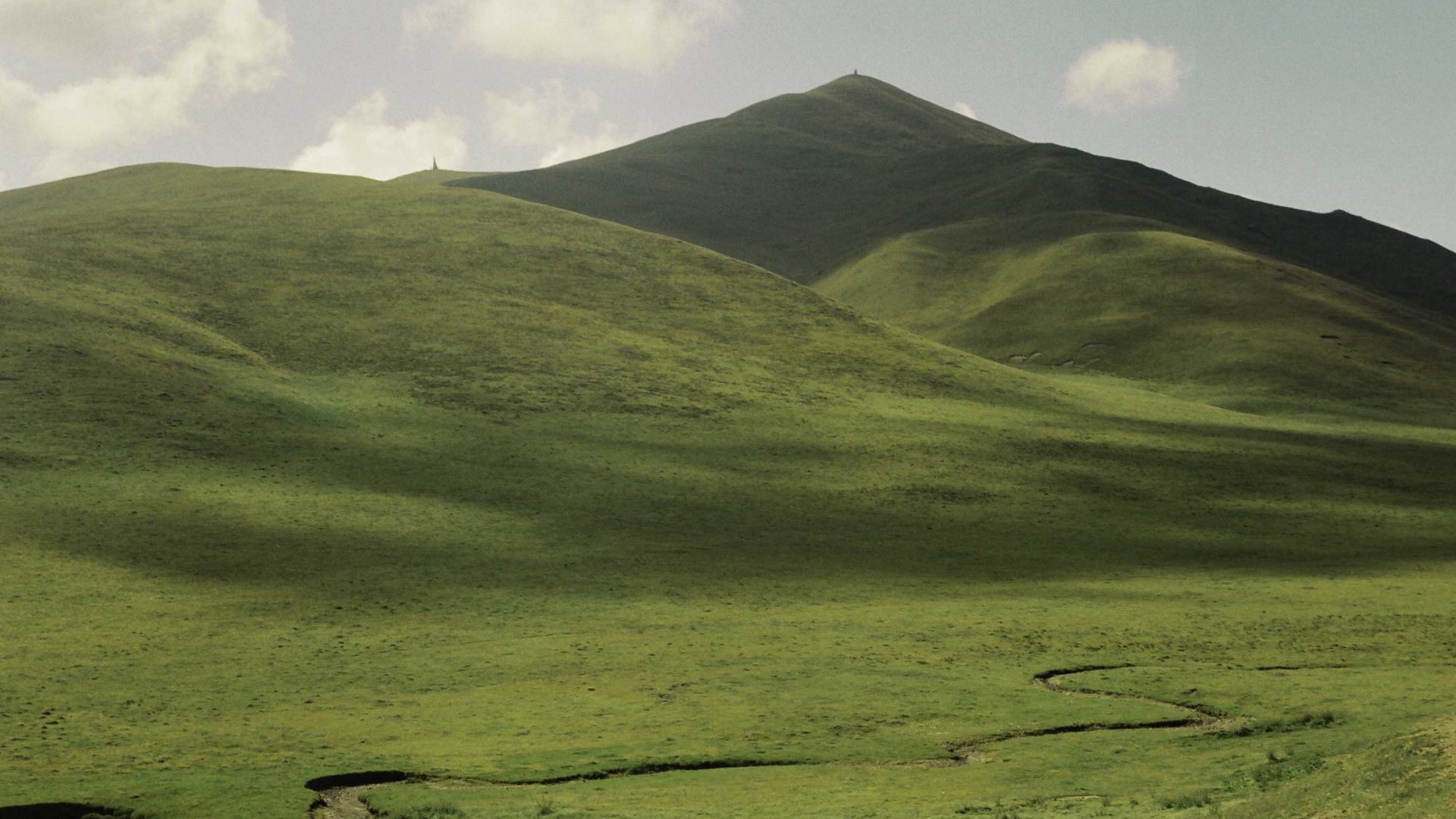 A path weaves through the green grass that leads to a similarly green mountain.